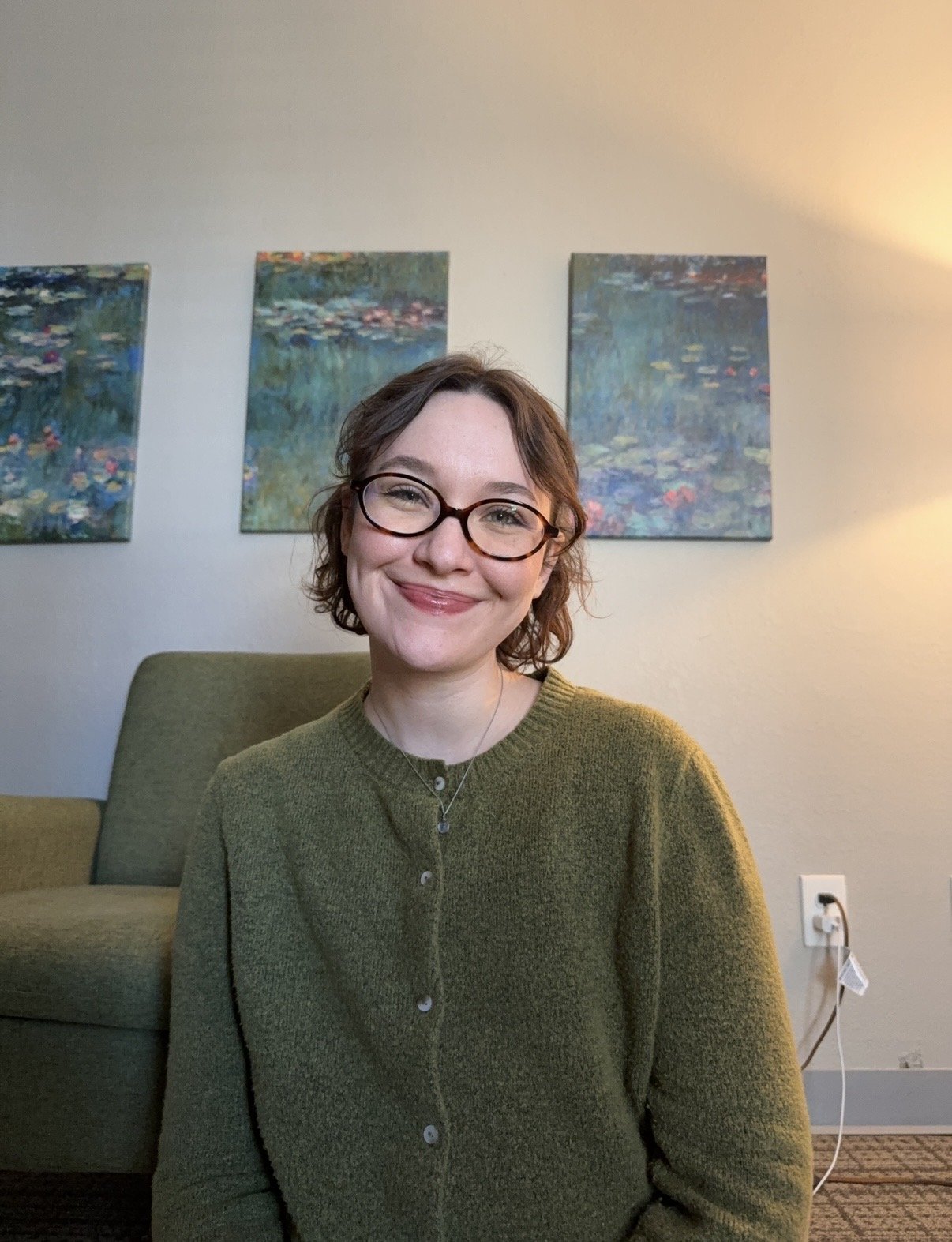 A woman with short curly brown hair, glasses, and a green cardigan smiling while sitting in a living room. Behind her are three paintings of water lilies on the wall and a green couch. An electrical outlet and plug are visible on the wall to her right.