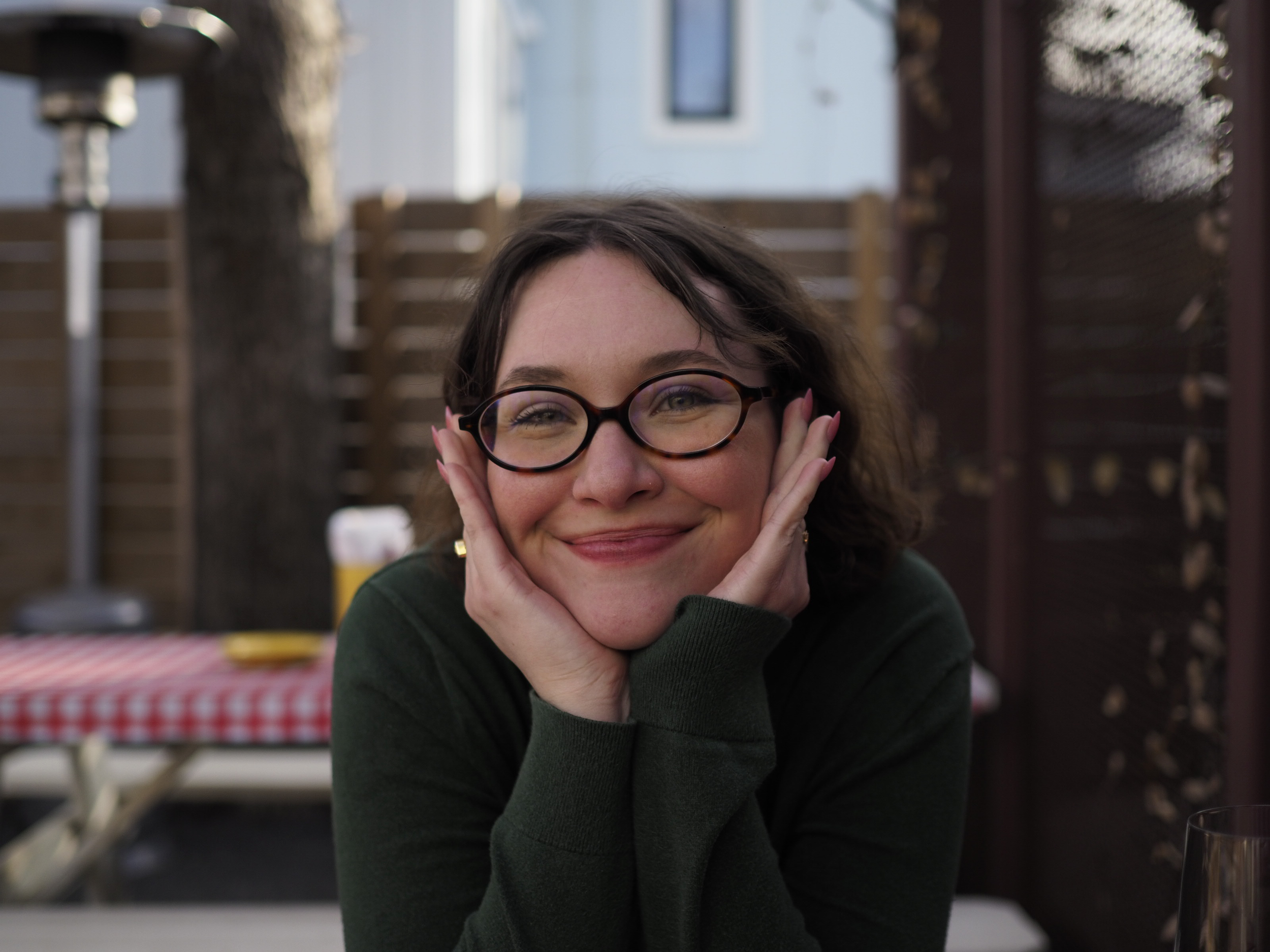 A woman with glasses smiling and resting her chin on her hands, sitting at a table outdoors with a tree, a fence, and a building in the background.
