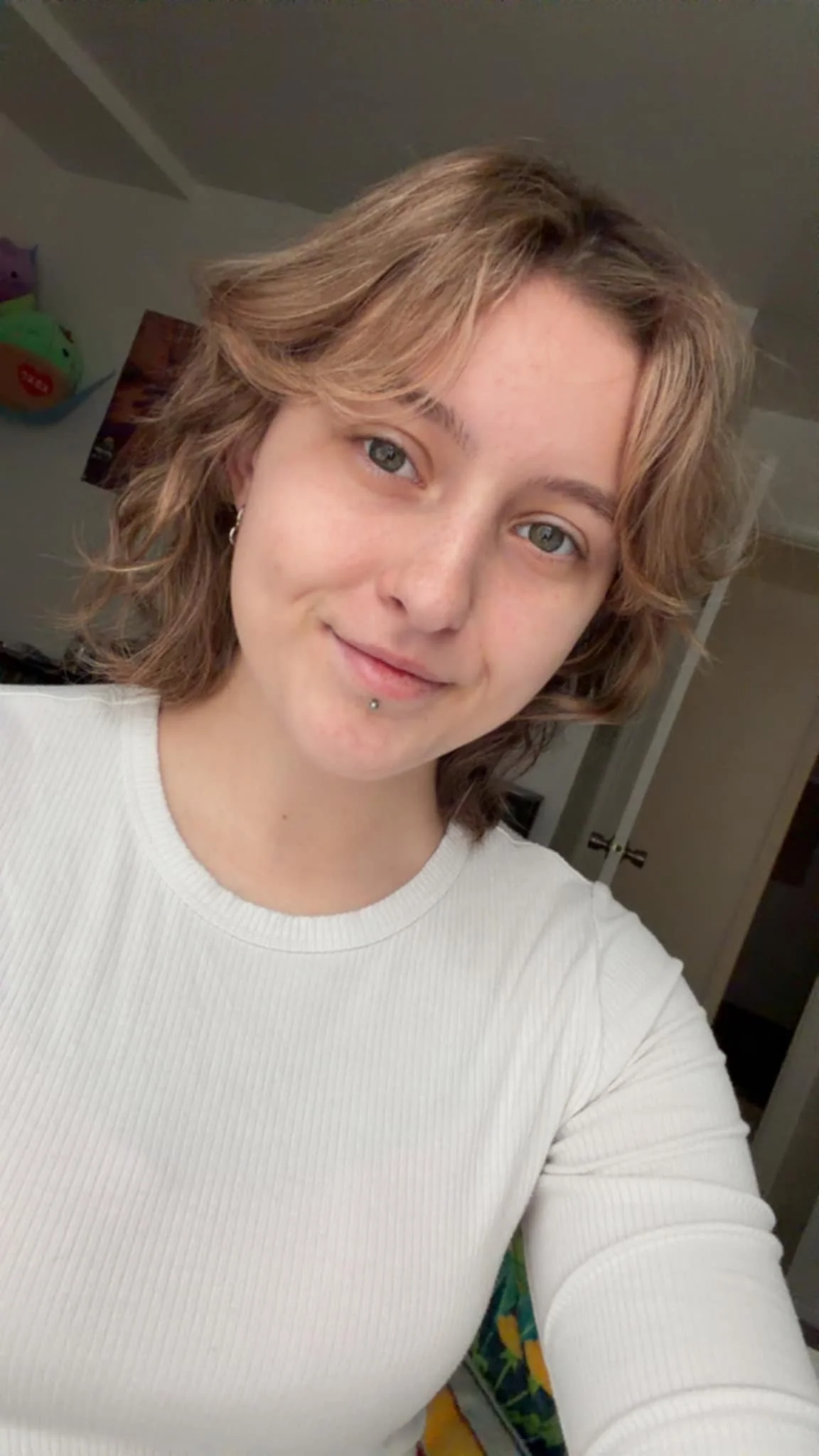 A young woman with short, wavy, light brown hair taking a selfie in a room, wearing a white long-sleeve shirt.