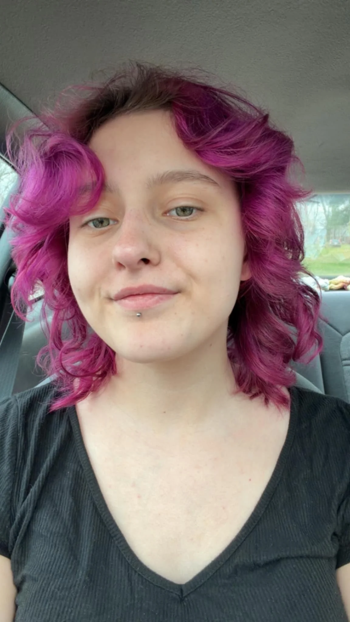 A young woman with short, wavy hair dyed vibrant purple-pink, sitting inside a car with a neutral expression.
