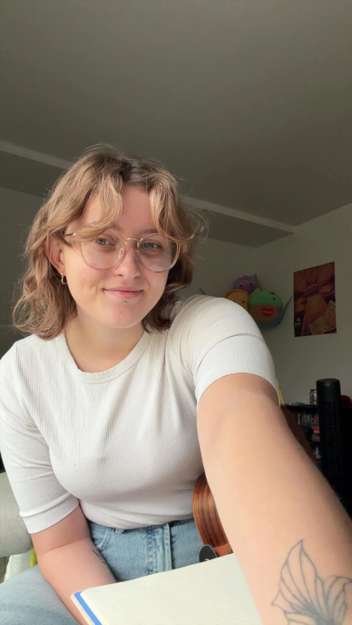 A young woman with glasses and piercings smiling while taking a selfie in a room with colorful plush toys and posters in the background.