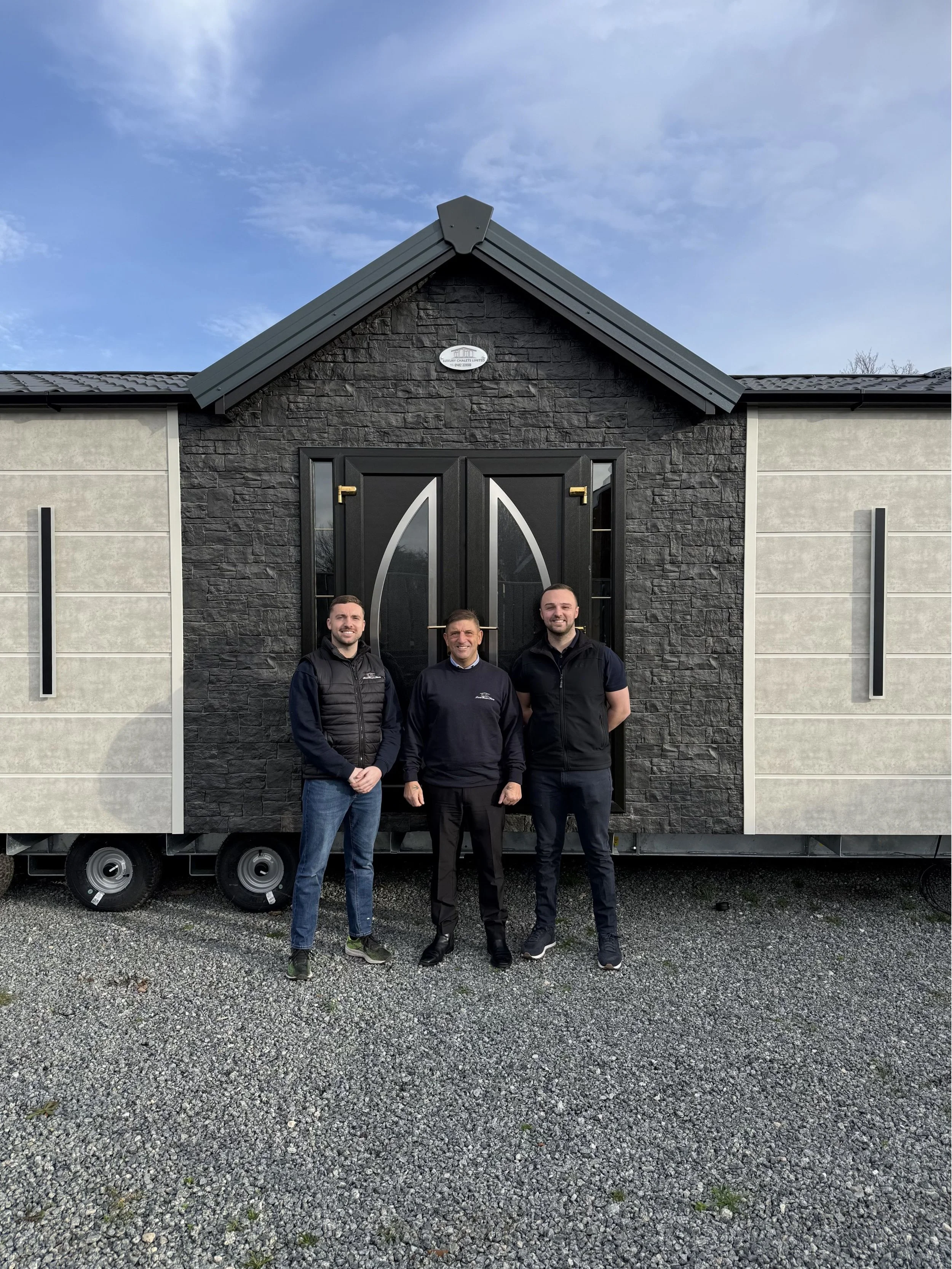 Three men standing outside in front of a small house on a trailer, with a gravel ground and a blue sky in the background.
