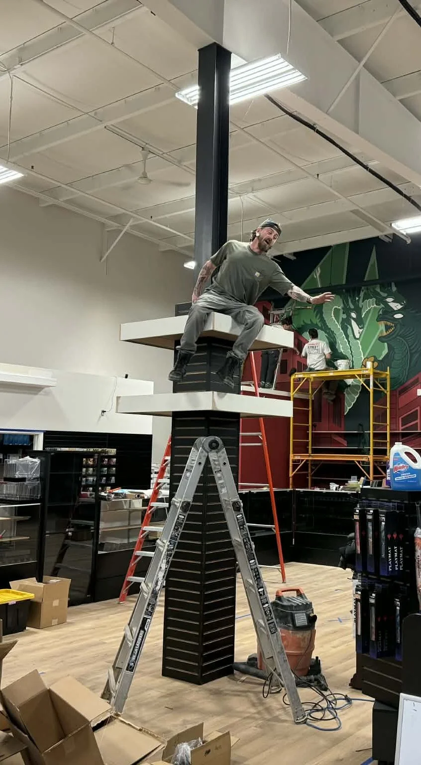 A man standing on a small platform on top of a column, appearing to lean and pose with a playful expression. The setting is an indoor space, possibly a store or workshop, with ladders and tools around.