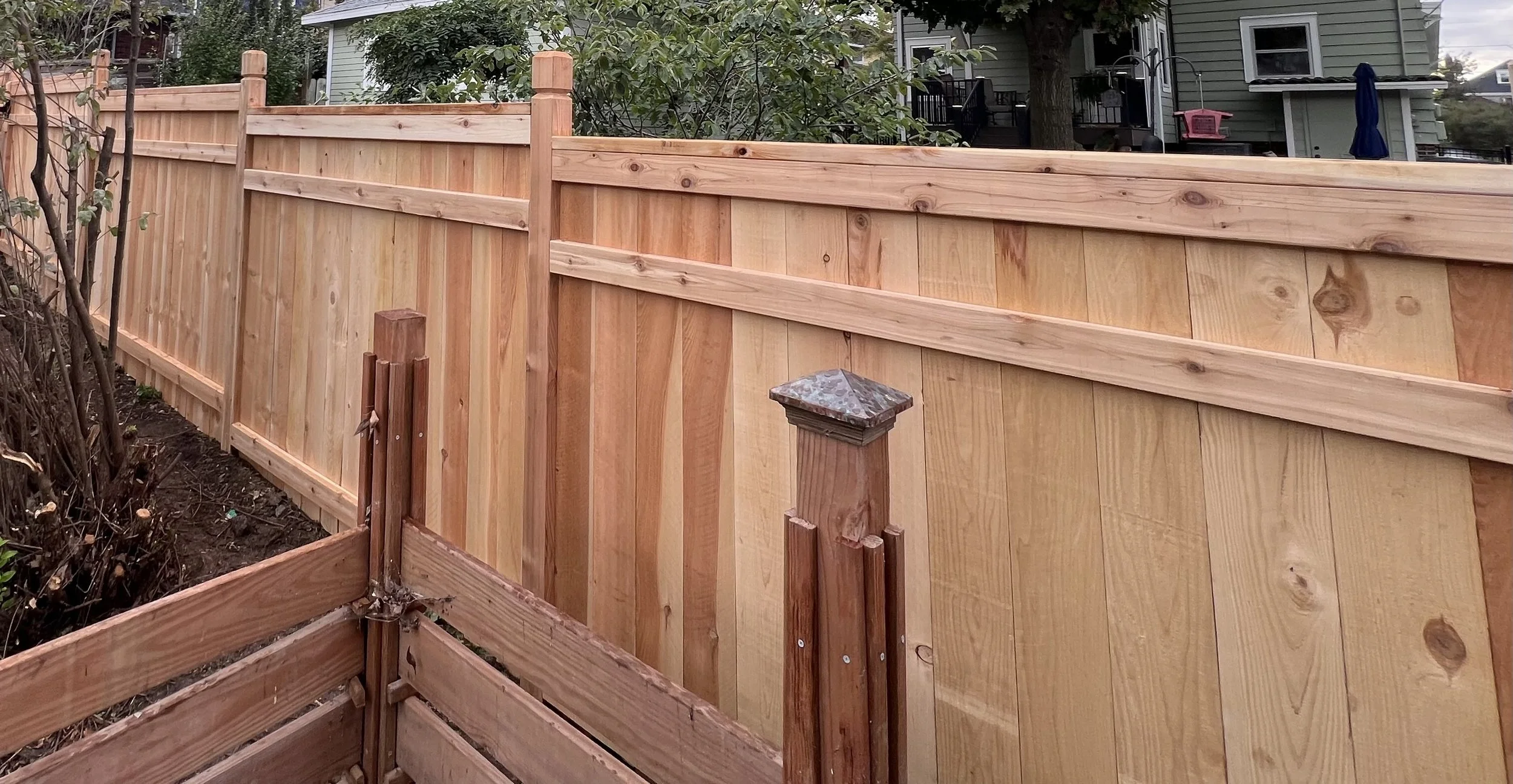 Newly constructed wooden privacy fence with vertical panels, supported by posts, surrounding a backyard with a house and trees in the background.