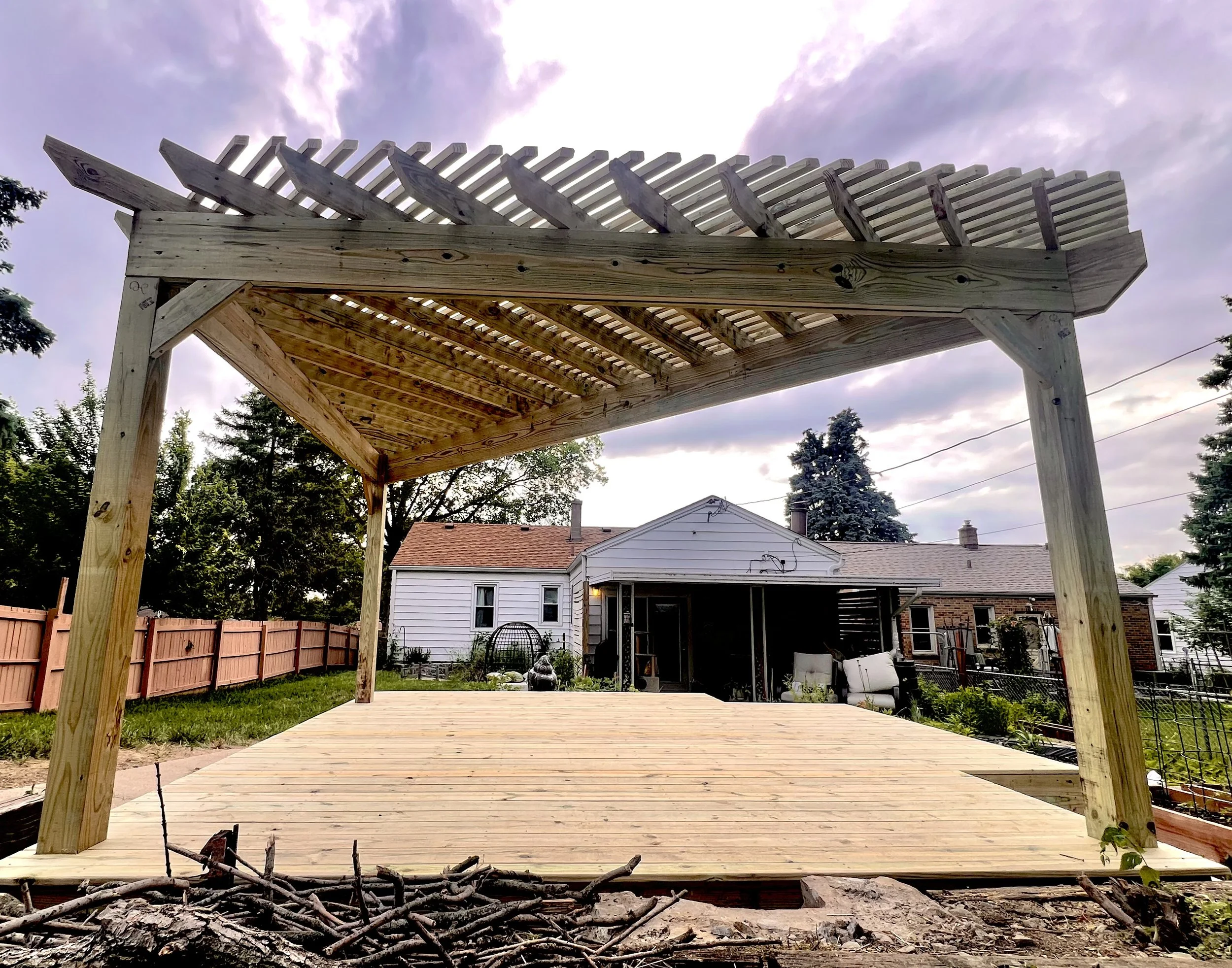 Newly built wooden deck with a pergola in a backyard, with houses and trees in the background under a cloudy sky.