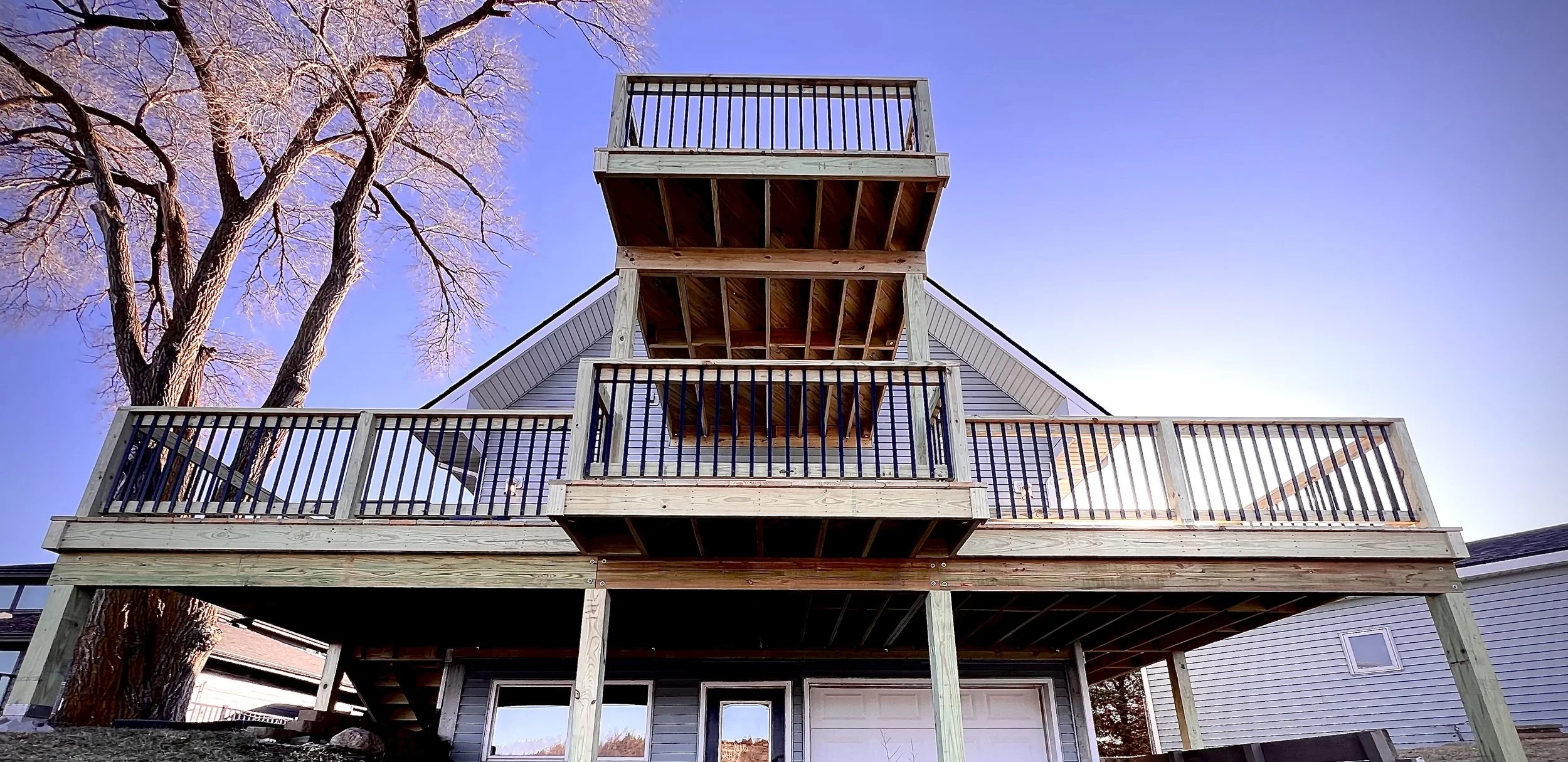 A three-tiered wooden deck structure attached to a house, with black railings and a large tree with leafless branches nearby, under a clear blue sky.