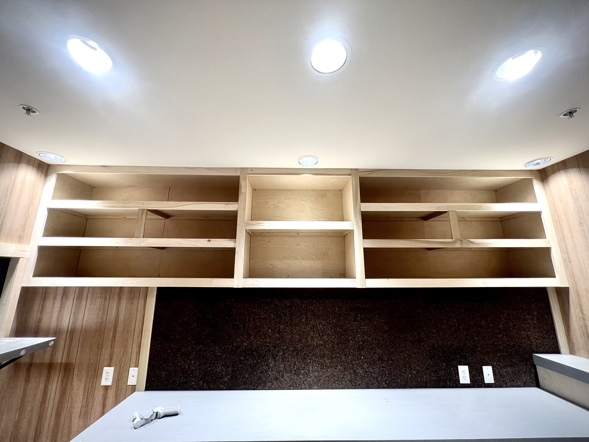 Empty wooden shelving unit above a countertop in a room with ceiling lights.