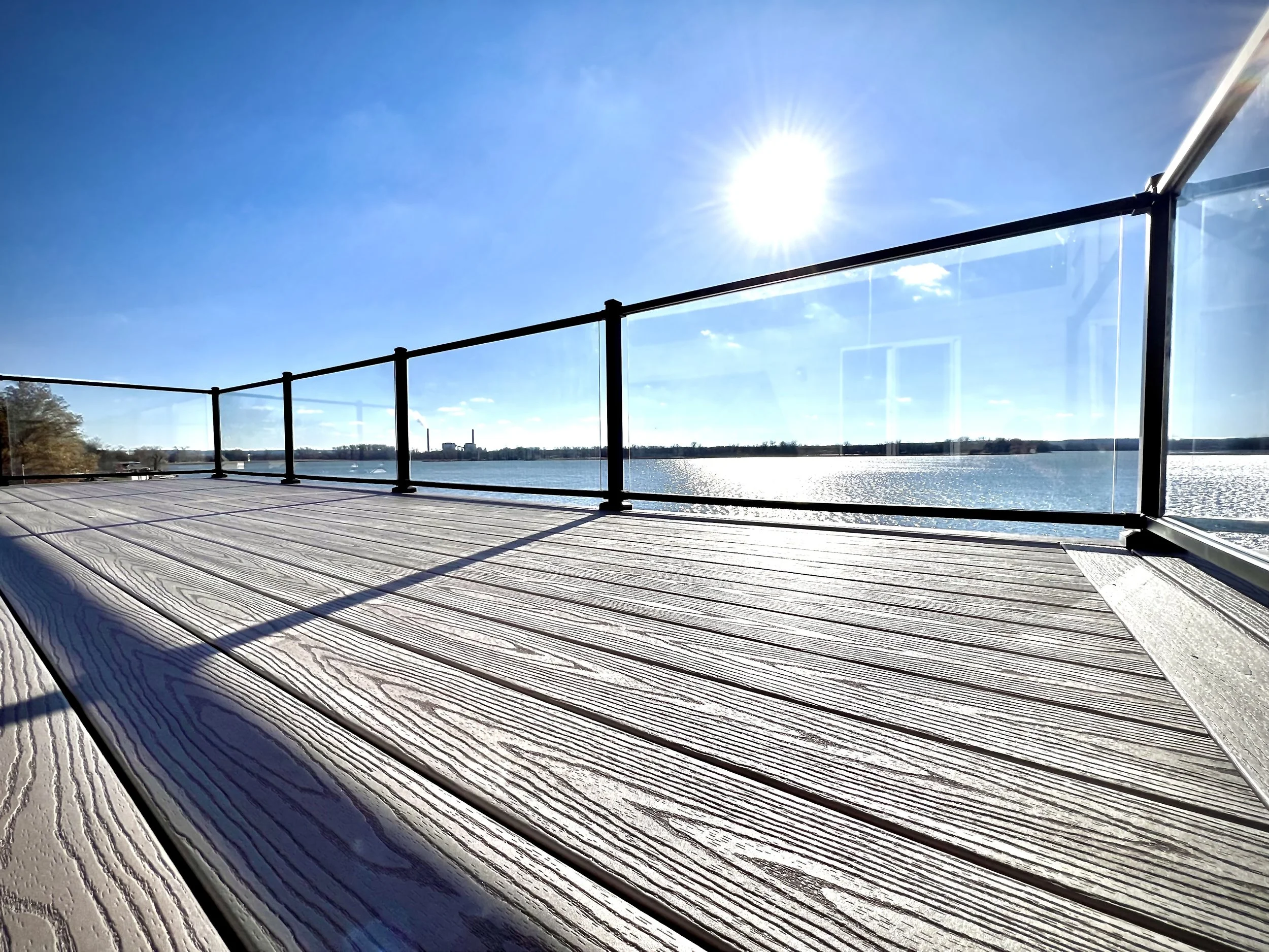 A wooden deck overlooking a body of water with a clear blue sky and the sun shining brightly overhead. The deck has glass railings with black posts, reflecting the sunlight.