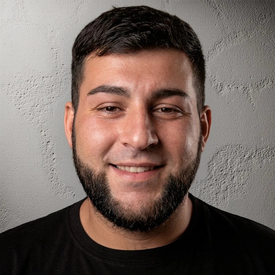 Retrato de un hombre con barba, sonriendo, cabello castaño oscuro, usando una camiseta negra, fondo gris con textura.