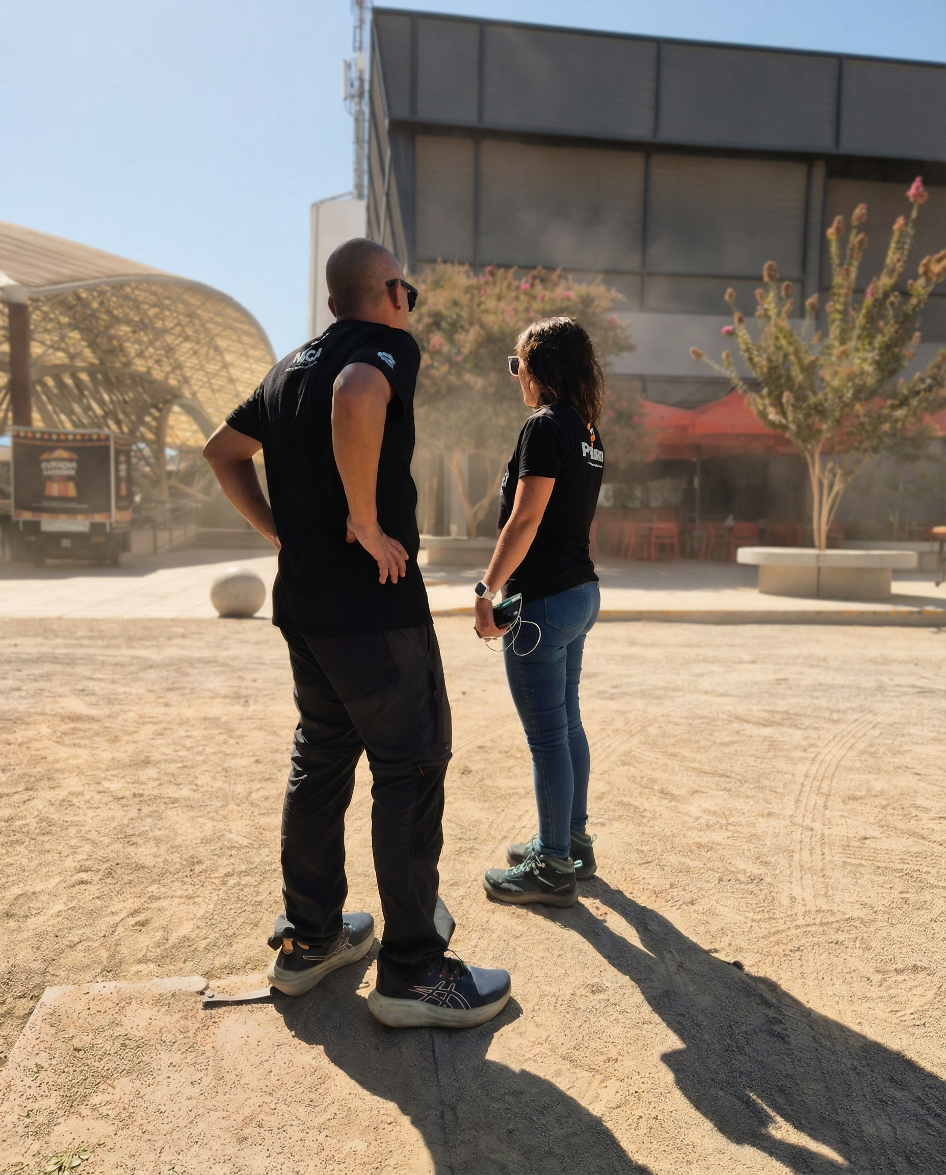 Dos personas con camisetas negras conversando en un espacio al aire libre, con edificios y árboles en el fondo.