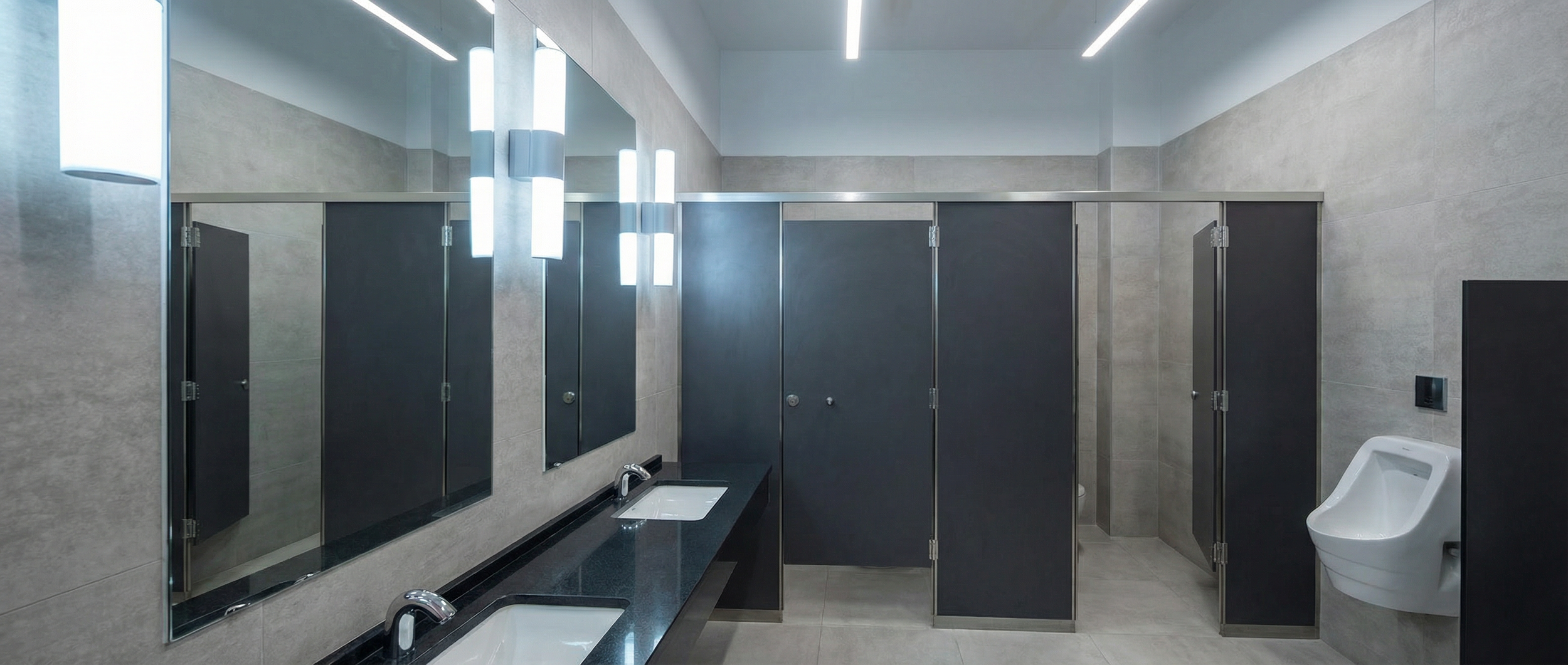 Lavabo y espejos en baño público con limitadores de agua, puertas de privacidad, iluminación moderna y un inodoro en cabina.