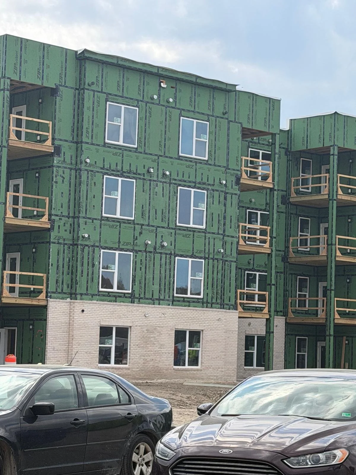 Under construction multi-story building with green sheathing, several windows, and wooden balconies, with cars parked in front.