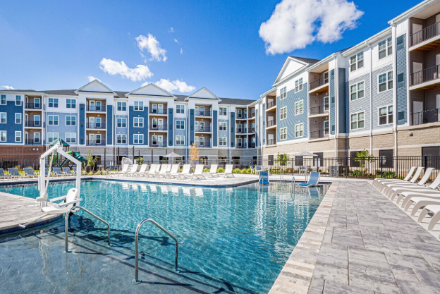 Outdoor swimming pool area in an apartment complex with lounge chairs, surrounded by a multi-story residential building under a partly cloudy sky.