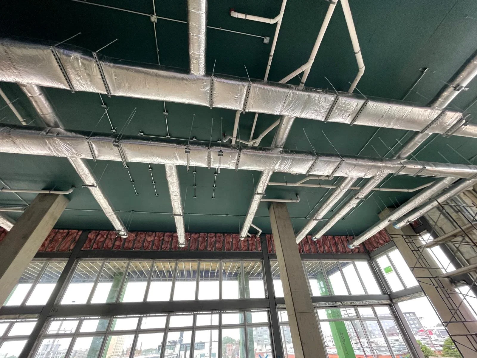 View of interior ceiling with exposed silver ventilation ducts and white piping, supported by metal brackets, against a dark green ceiling, with large windows at the bottom revealing an urban cityscape outside.