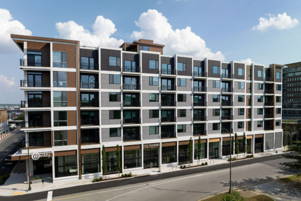 Modern multi-story residential building with retail shops on the ground floor, large windows, and a street in the foreground.