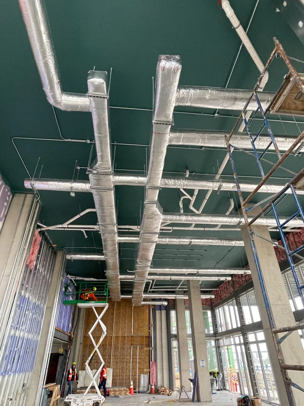 Construction workers installing ductwork in a building's ceiling, with scaffolding and metal framework visible.