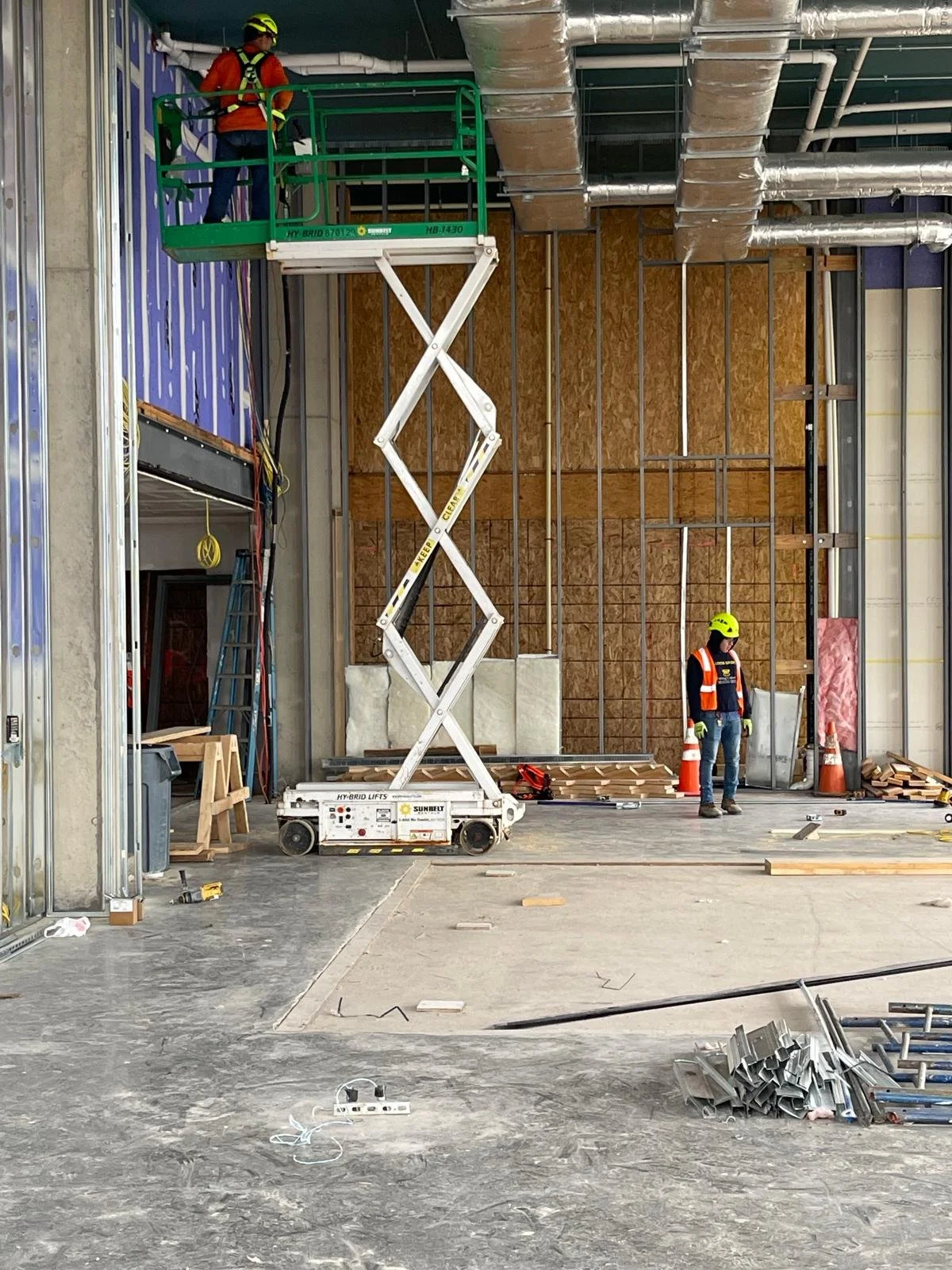 Construction workers using a scissor lift and standing on the ground inside a building under construction with exposed ductwork and unfinished walls.