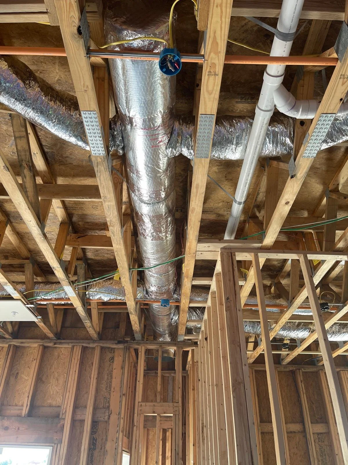 Construction site with exposed wooden framing, silver insulated ductwork, and white plumbing pipes in the ceiling area.