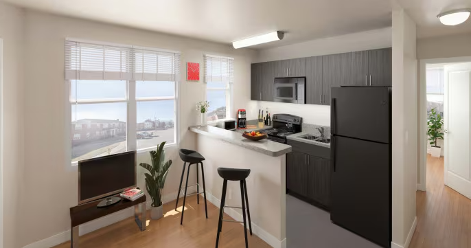 Modern kitchen with black appliances, dark wood cabinets, white countertops, and a breakfast bar with two black stools. Large windows with blinds overlook a neighborhood. There is a small TV, a plant, and some kitchen essentials on the counter.