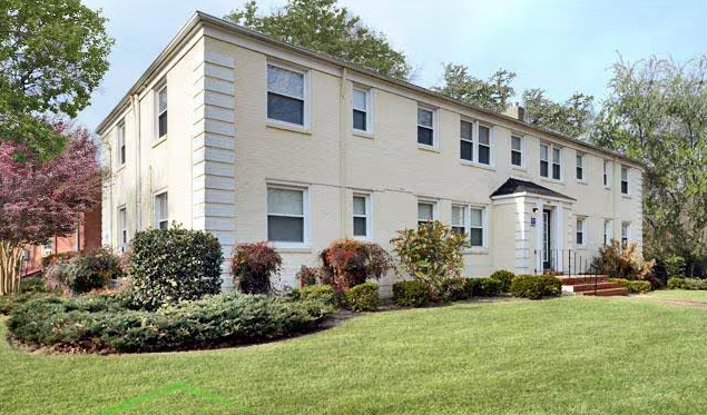 A white multi-family residential building with multiple windows and a small front porch, surrounded by landscaped bushes and a green lawn, with trees in the background.