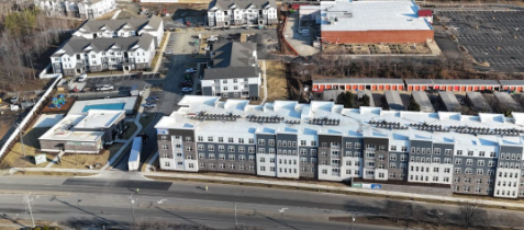 Aerial view of a modern apartment complex with white and gray buildings, surrounded by parking lots and nearby streets.