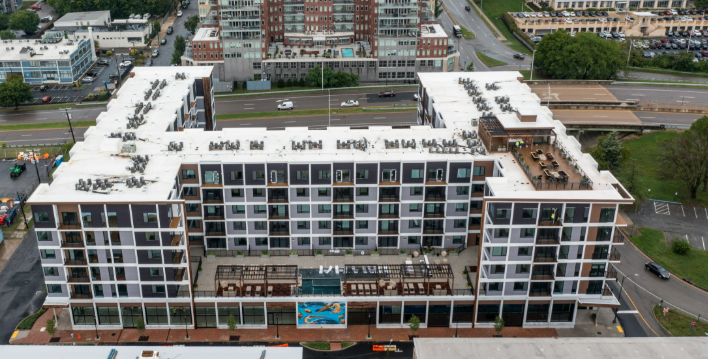Aerial view of a modern, white multi-story apartment building with outdoor amenities including a swimming pool, located in an urban area with nearby roads and other buildings.