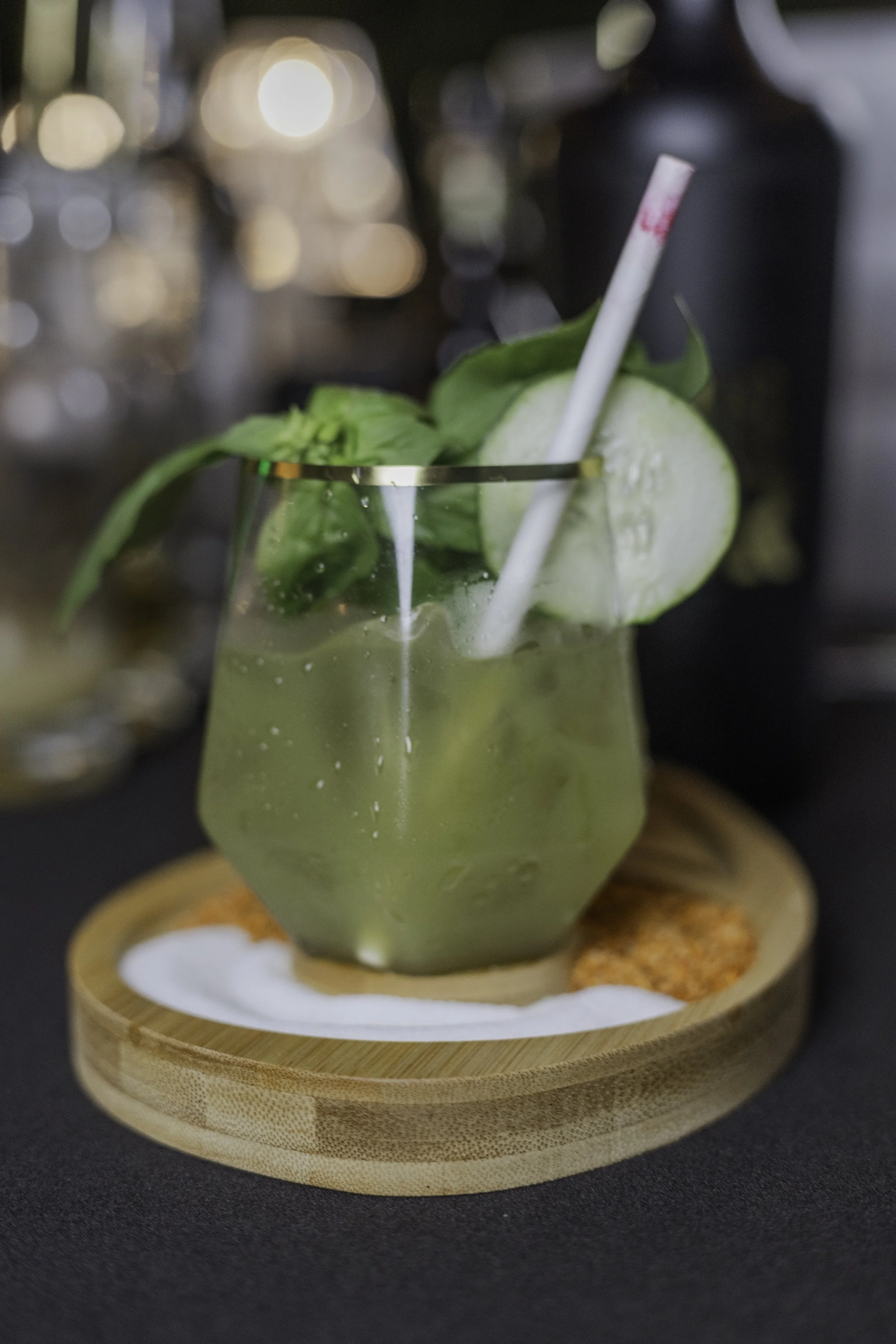 A drink with a slice of cucumber and fresh green mint leaves, served in a glass with a straw, on a wooden coaster with snacks around it.