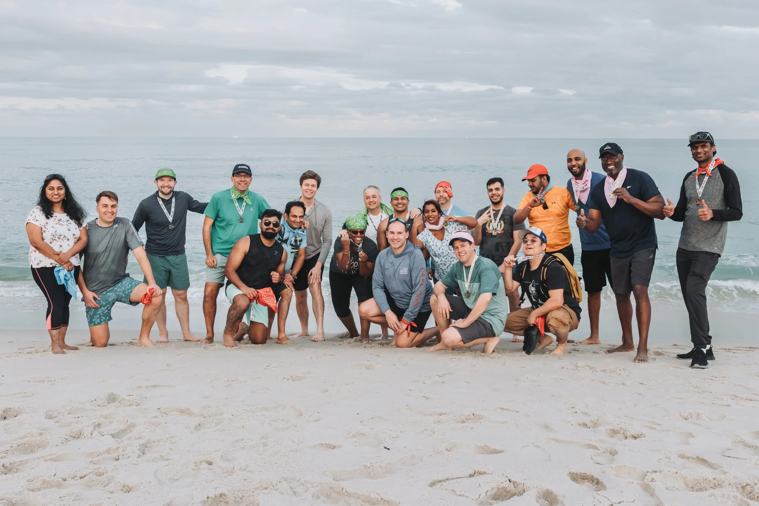 Group of 18 people standing and kneeling on a beach, smiling and posing for a photo, with the ocean and cloudy sky in the background.