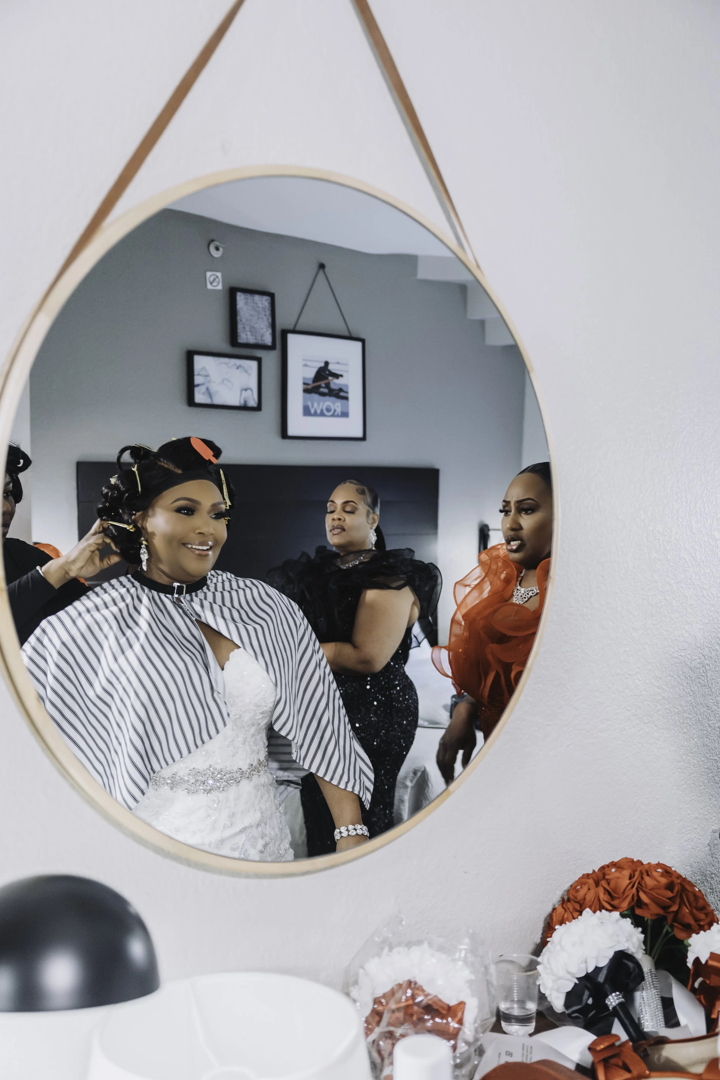 Women getting ready in front of a mirror, with one woman smiling and another appearing serious. The reflection shows hairstyles and clothing, with hair rollers and accessories visible. The setting is a room with framed pictures on the wall and a tabl