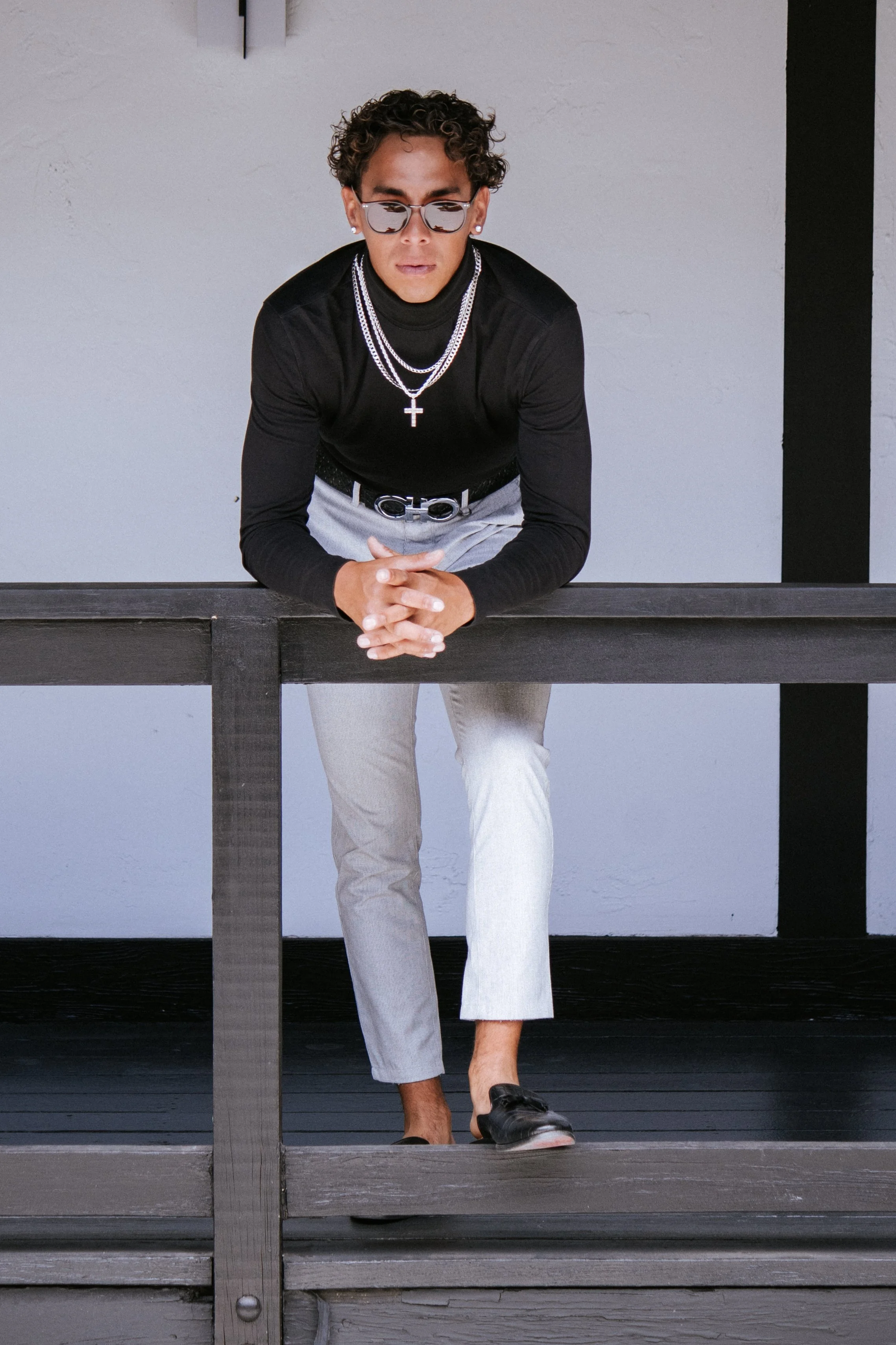 A person with curly hair and sunglasses, wearing a black long-sleeve shirt, layered necklaces, light-colored pants, and black shoes, leaning on a black wooden railing against a white wall with black vertical accents.