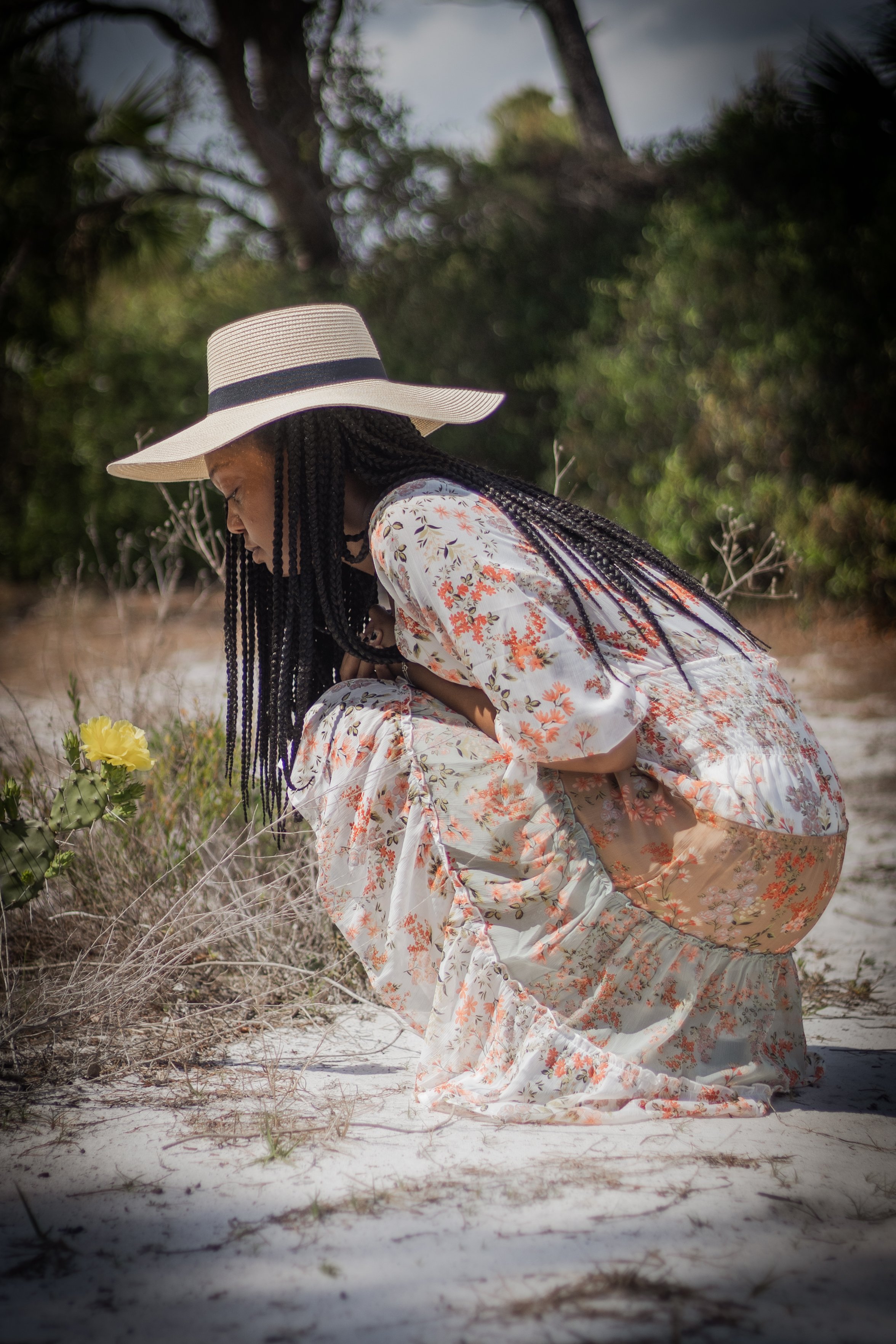 A woman with braided hair in a floral dress with orange and white patterns, wearing a large sun hat with black ribbon, is outdoors kneeling on the sandy ground near some yellow flowers and dry bushes, with trees in the background.
