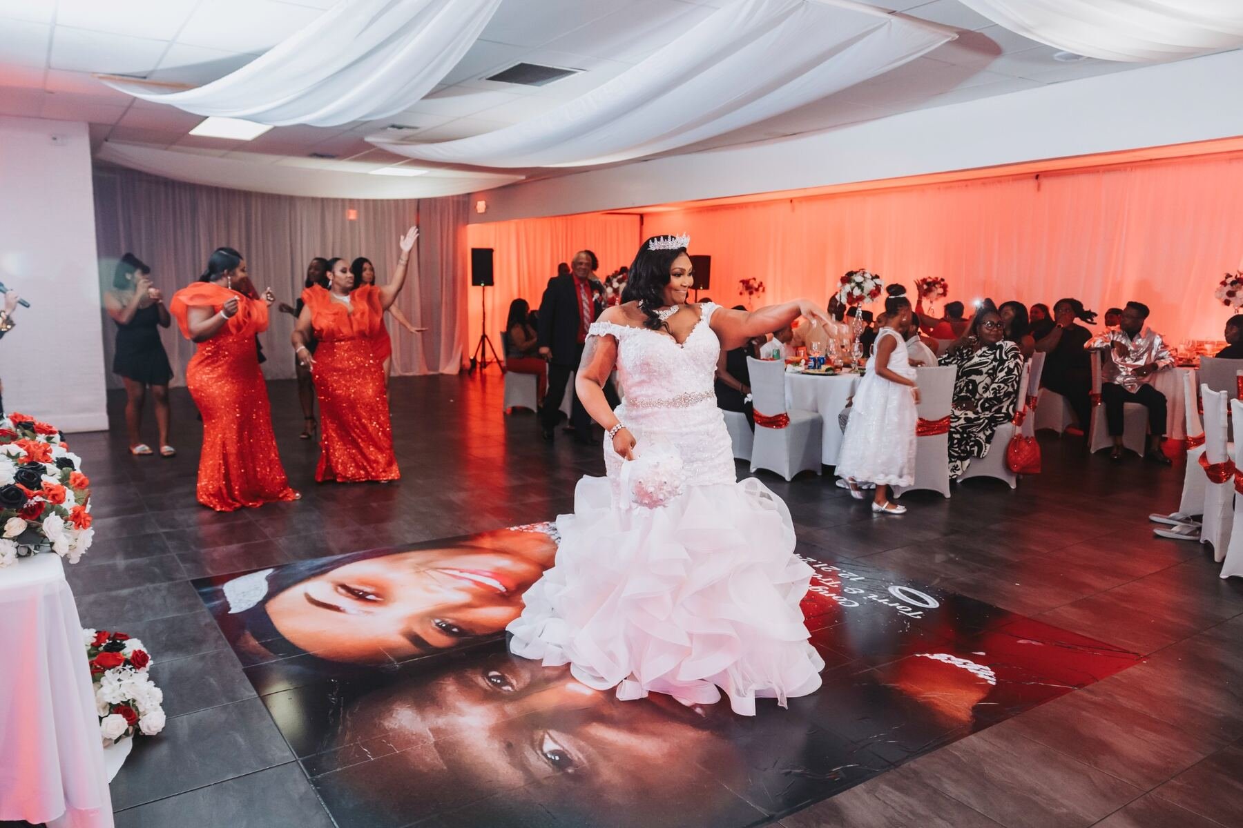 A bride in a white wedding dress with a tiara is smiling and dancing at a reception with guests watching. Guests are seated at decorated tables with red and white flowers. The dance floor has a large image of a woman's face