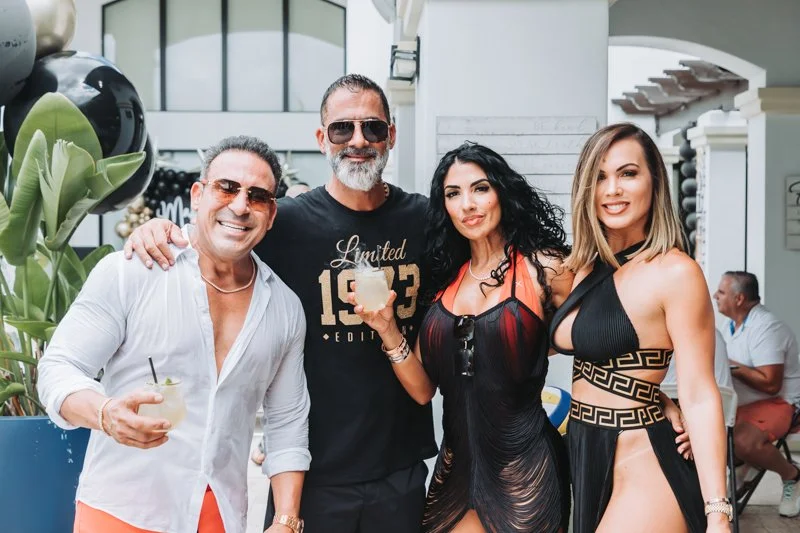 Group of four adults smiling and enjoying drinks at an outdoor party or event, with a modern building and other guests in the background.