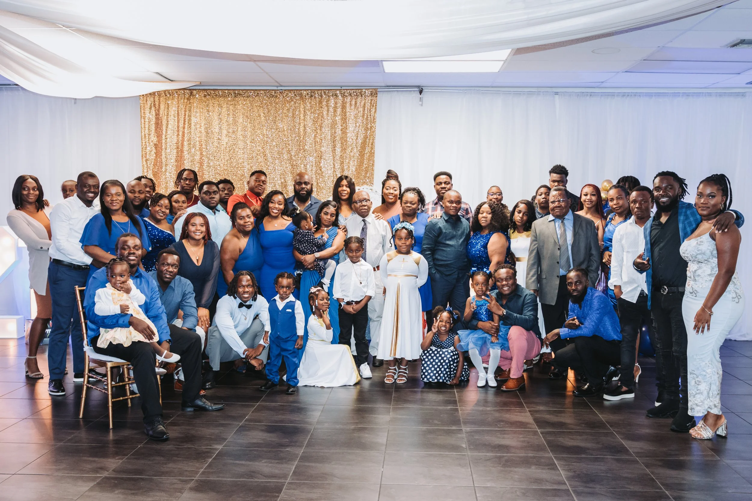 A large group of diverse people, including children, adults, and seniors, pose for a photo at a celebration or family gathering in a decorated indoor venue with white and gold accents. Many are dressed in blue, white, and formal attire.
