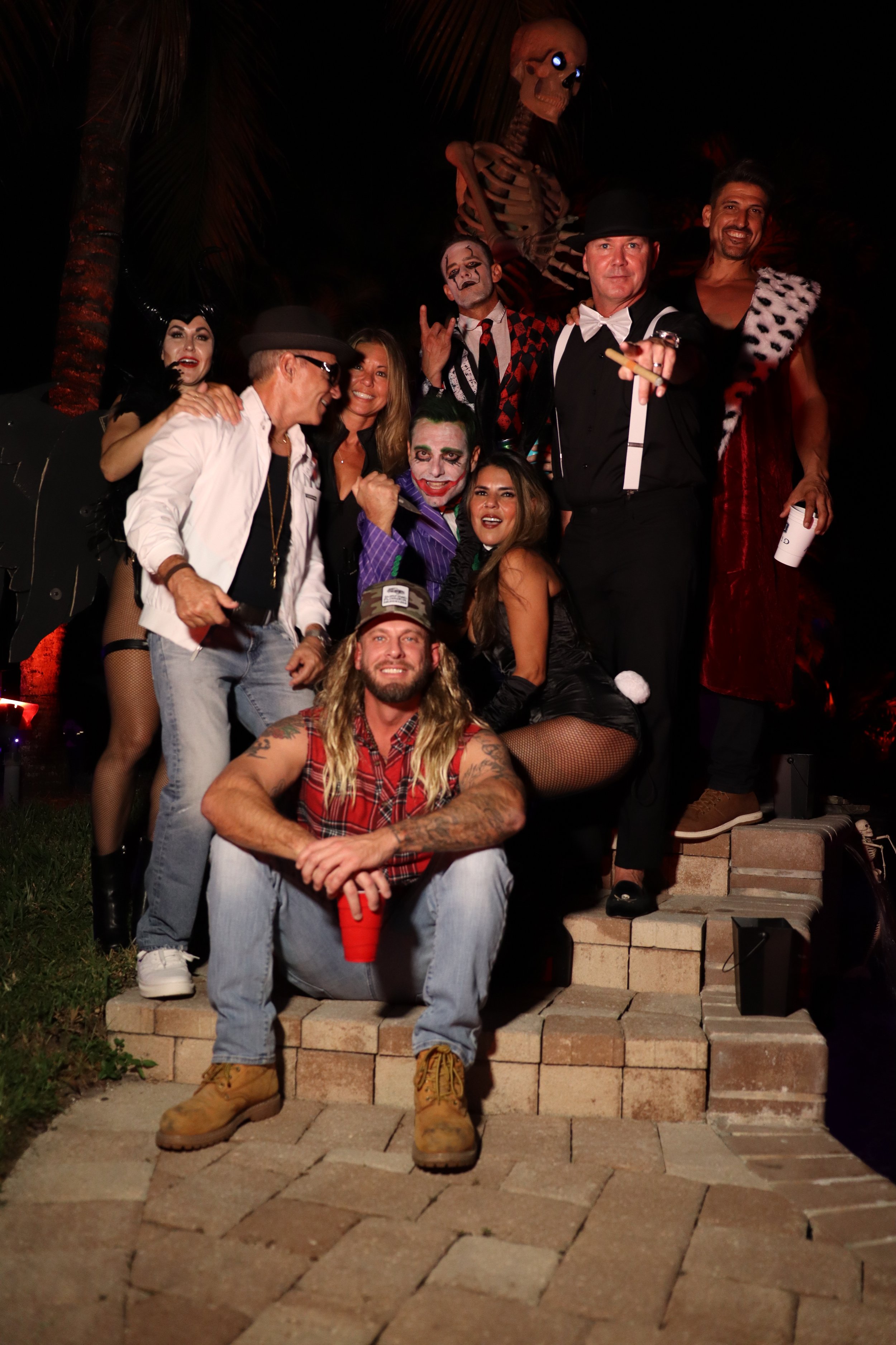 Group of people dressed in costumes, including clowns and characters, standing on stairs outdoors at night, some smiling and posing for the photo, with a skeleton prop and Halloween decorations in the background.