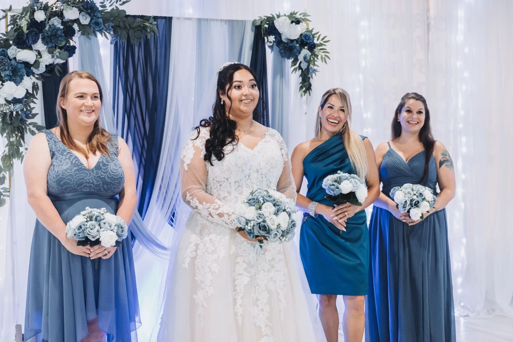 A bride and three bridesmaids standing in front of a blue and white floral backdrop, each holding a bouquet of white and blue roses. The bride is in a lace wedding dress, smiling, with dark hair and a tiara. The bridesmaids are in blue dresses, smili