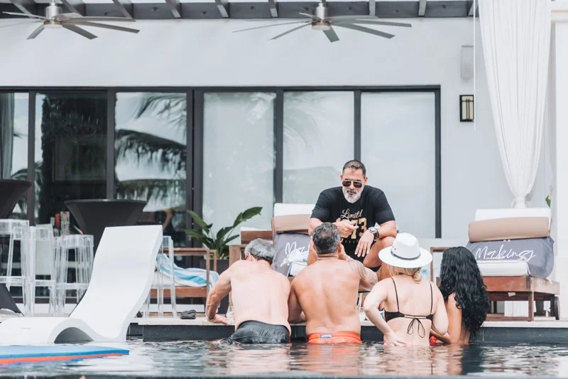 Multiple people in swimwear gathering in a pool area with a man sitting on a deck at a table, behind large glass sliding doors and white curtains.