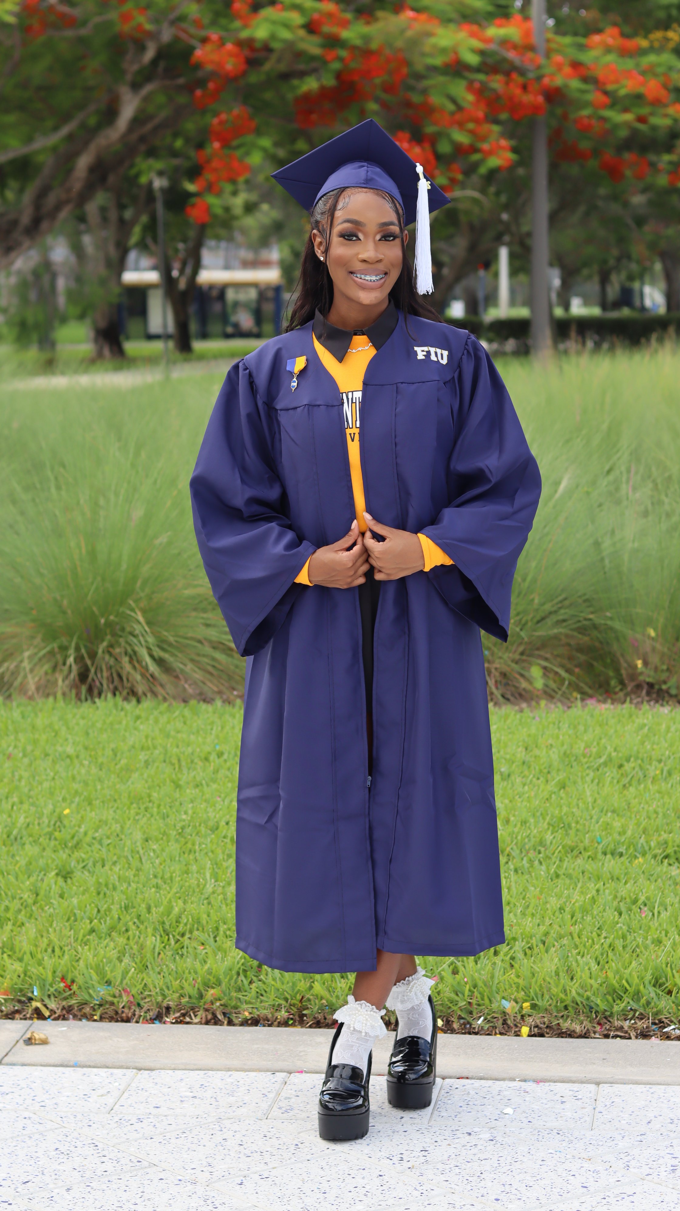Young woman in a navy graduation gown and cap standing outdoors on a sidewalk with green grass and trees with red flowers in the background, smiling and looking at the camera.