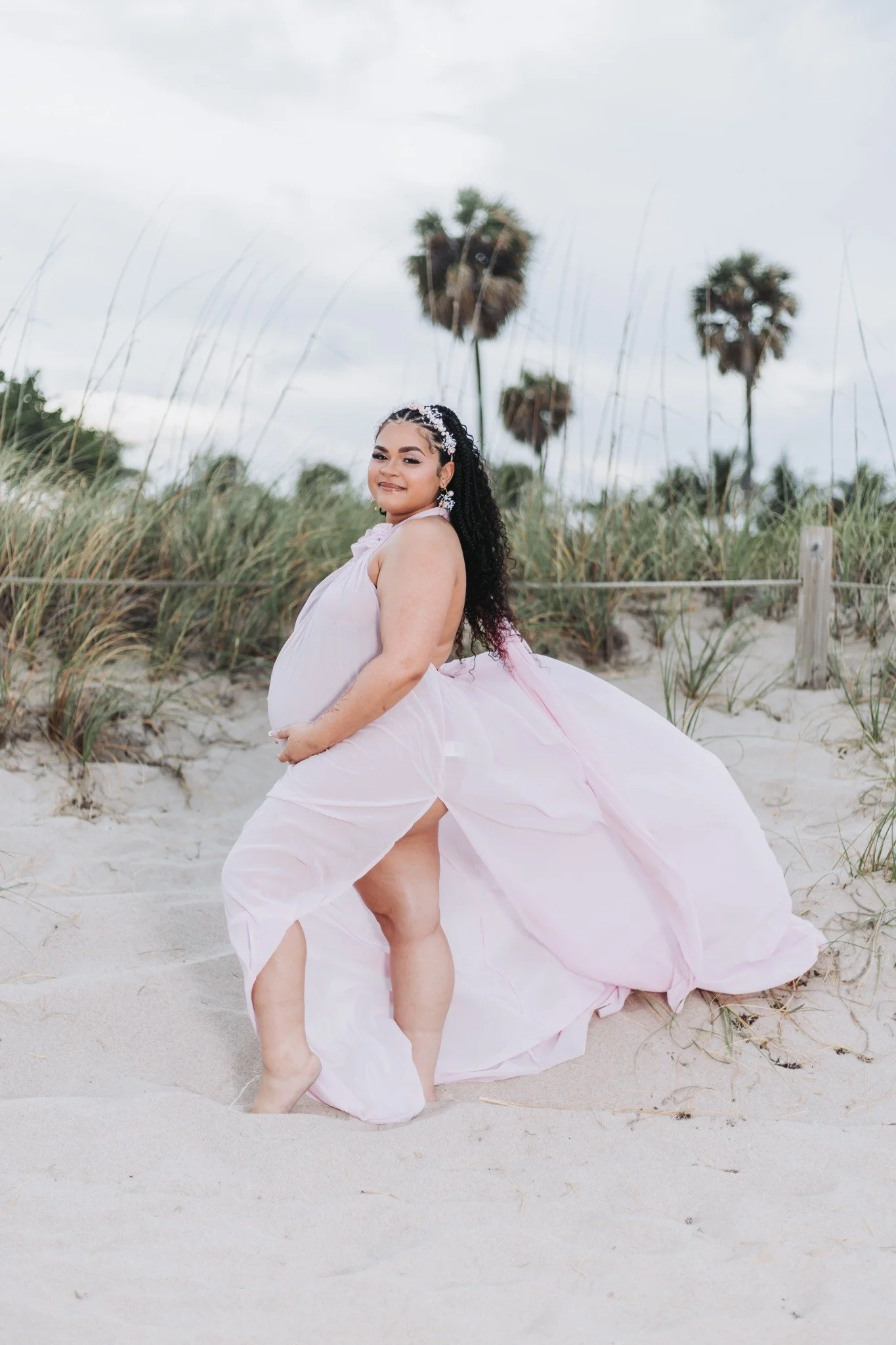 A pregnant woman dressed in a flowing pink dress standing barefoot on the sand at the beach, with tall grasses and palm trees in the background, outdoor cloudy sky.