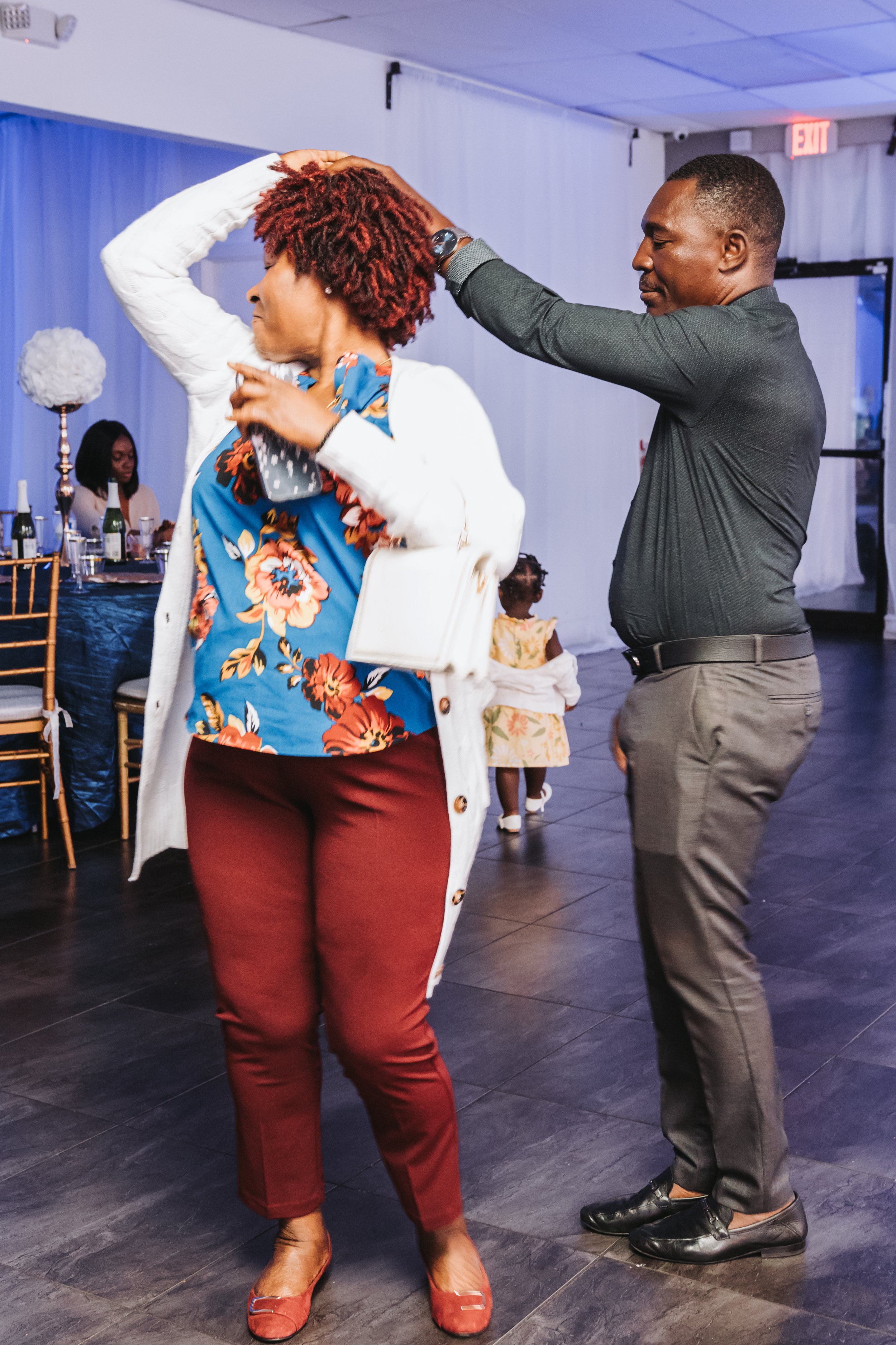 A man and woman dancing together in a formal setting, with other guests seated at tables in the background. The woman has red curly hair, wearing a floral blouse, white jacket, and maroon pants, while the man is wearing a gray shirt, gray pants, and 