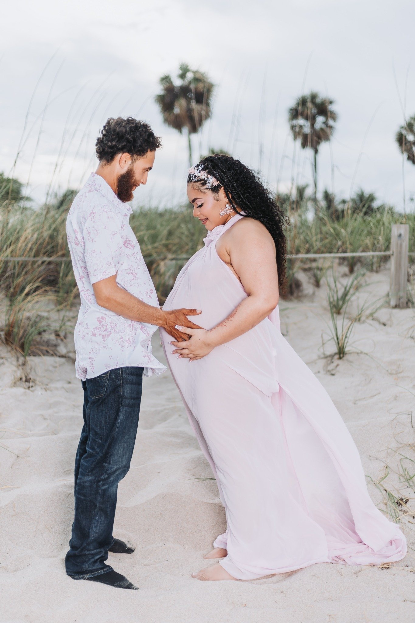 A couple on the beach, the man with curly dark hair and beard, wearing a light pink shirt and jeans, and the pregnant woman with long dark curly hair, wearing a flowing pink dress and floral hair accessories, standing close together with their forehe