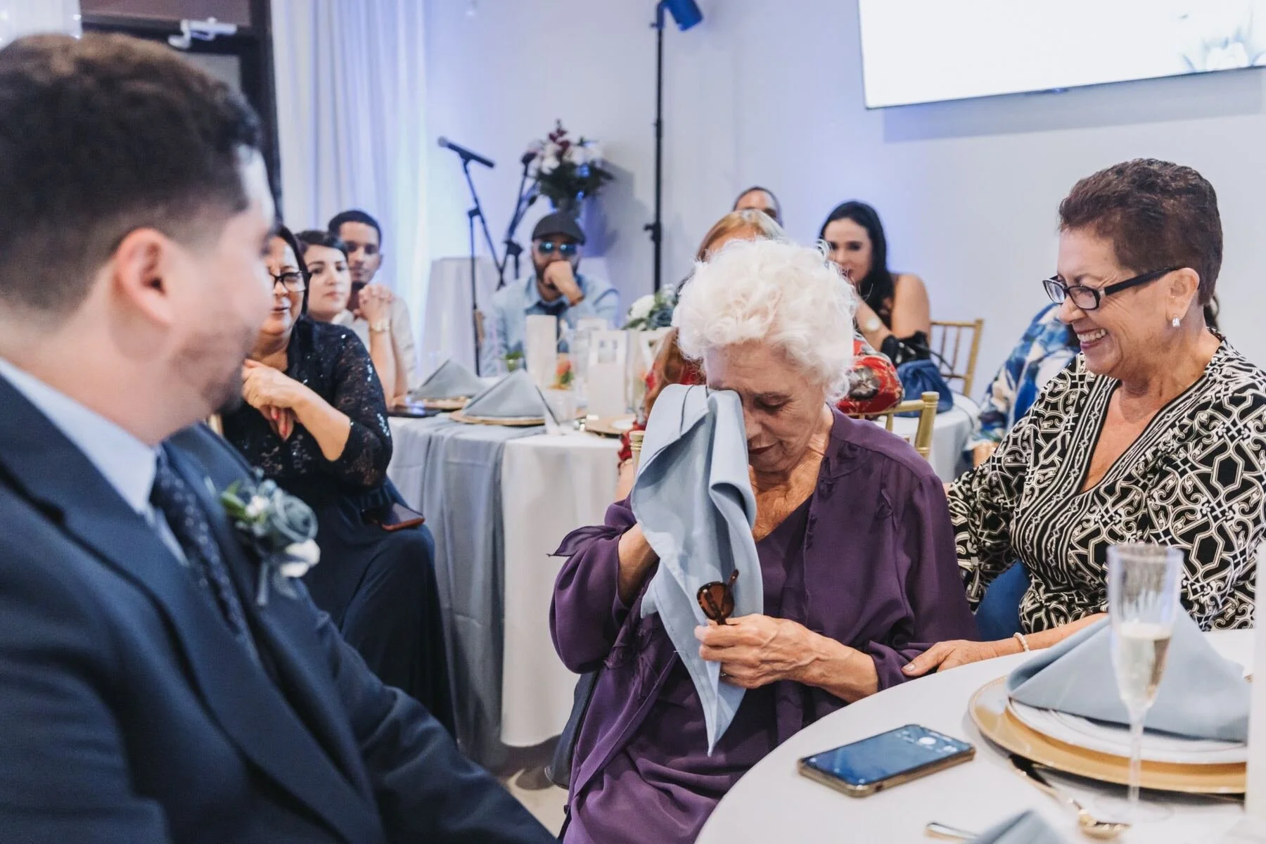 Guests seated at a formal event with a woman wiping tears from her eyes, a smiling woman next to her, and others observing the scene, during a joyful moment.