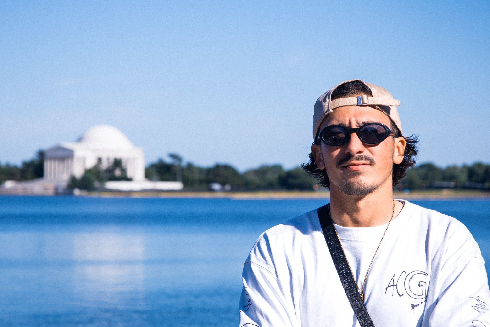 A young man wearing sunglasses, a beige cap backwards, a white shirt, and a camera strap around his neck standing outdoors near water with the Washington Monument in the background.