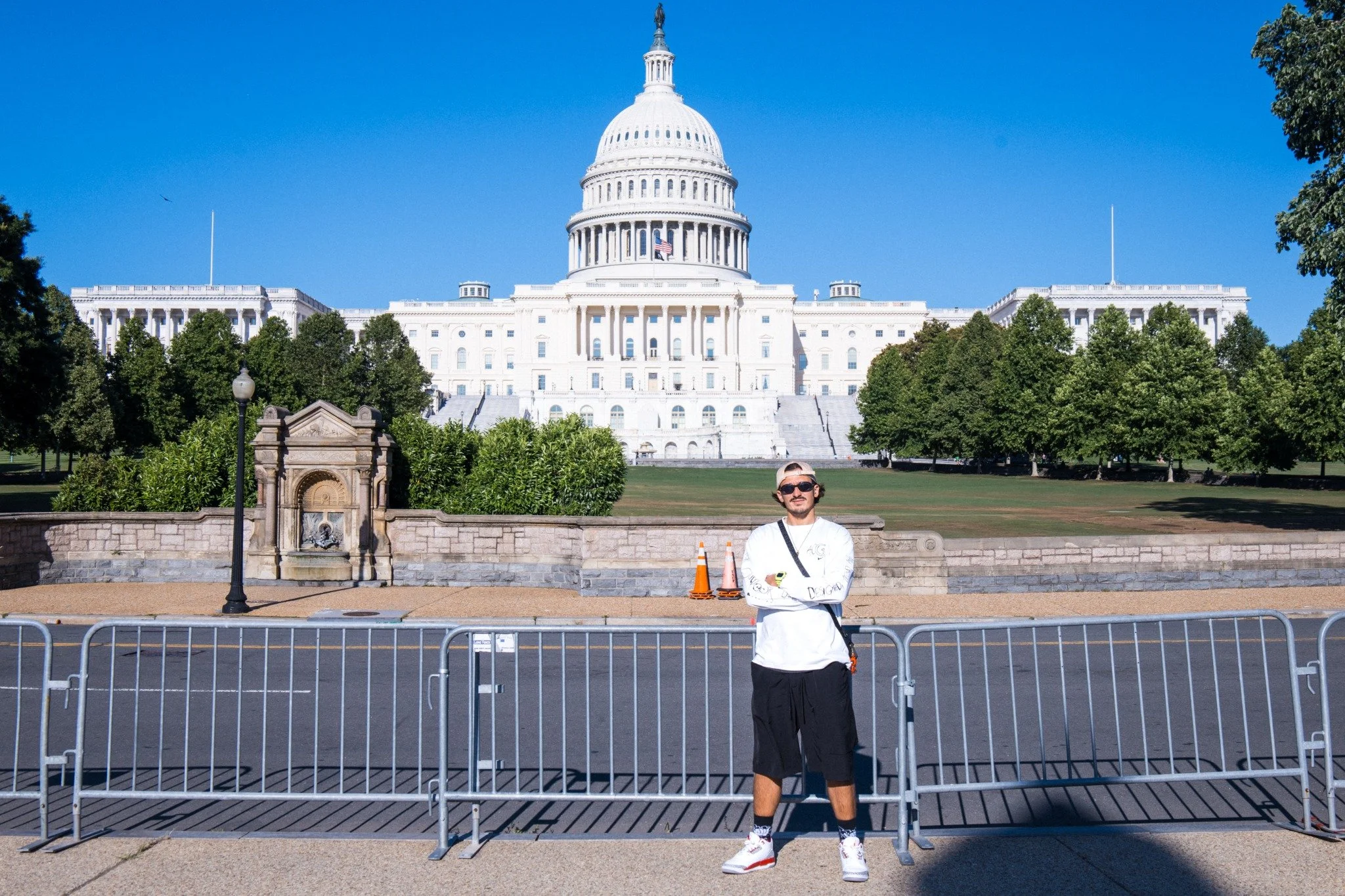 A man wearing sunglasses, a white long-sleeve shirt, and black shorts standing in front of the U.S. Capitol building with a clear blue sky, trees, and a fountain behind him. He is holding a black bag across his chest and posing with his arms crossed.