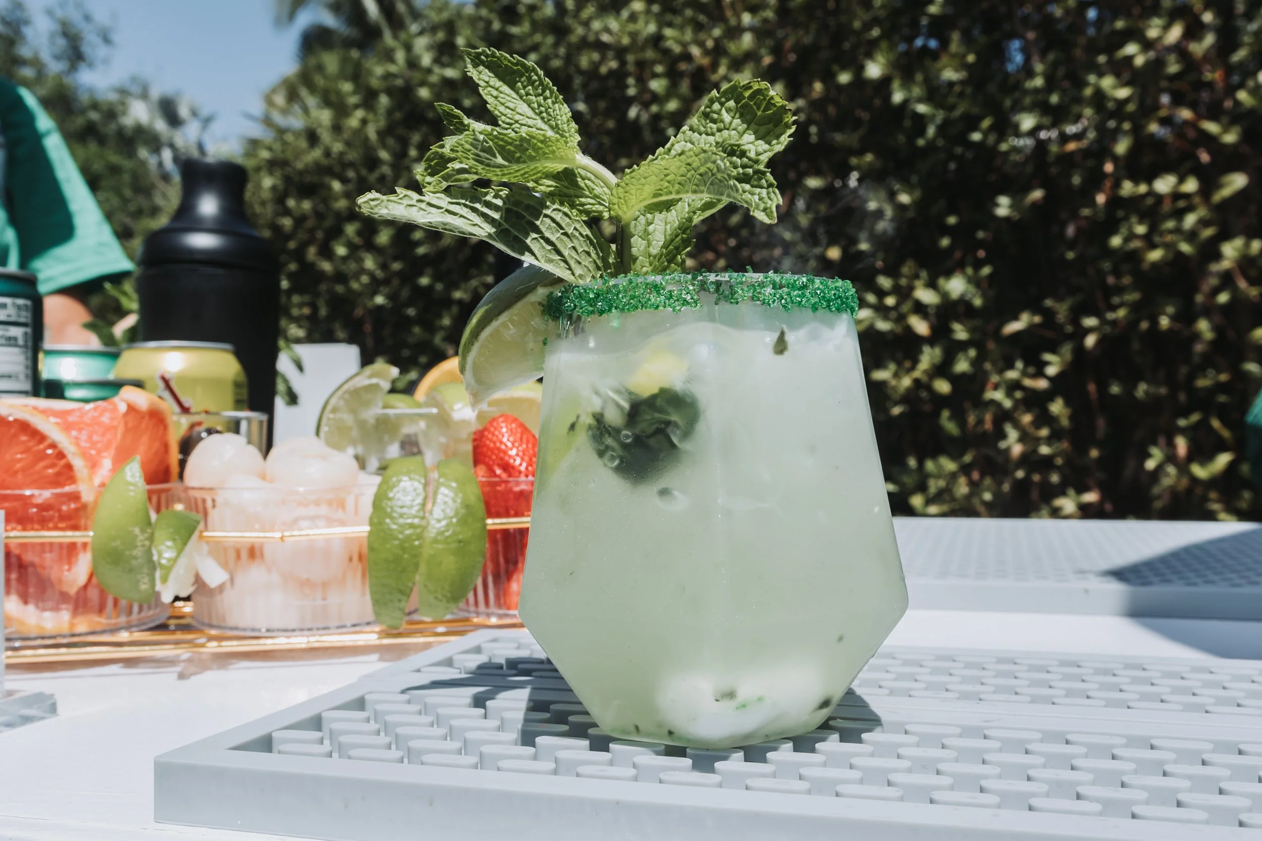 A refreshing cocktail garnished with fresh mint leaves, lime wedges, and green sugar on the rim, set on a white lattice table outdoors with a blurred background of trees and a table of various sliced citrus fruits.