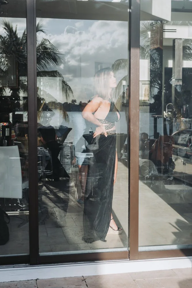 A woman dressed in a black evening gown with high slit, standing behind a glass window, with reflections of palm trees, water, and outdoor scenery visible on the glass.