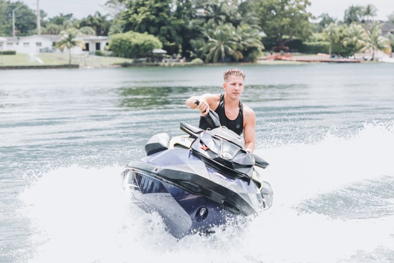 A young man riding a black jet ski on a lake, with trees and houses visible in the background.