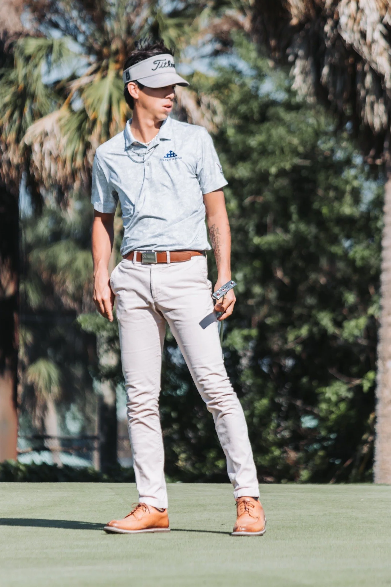 A person practicing golf on a green, wearing a light-colored golf shirt, beige pants, brown shoes, and a golf visor, holding a golf club, with trees in the background.