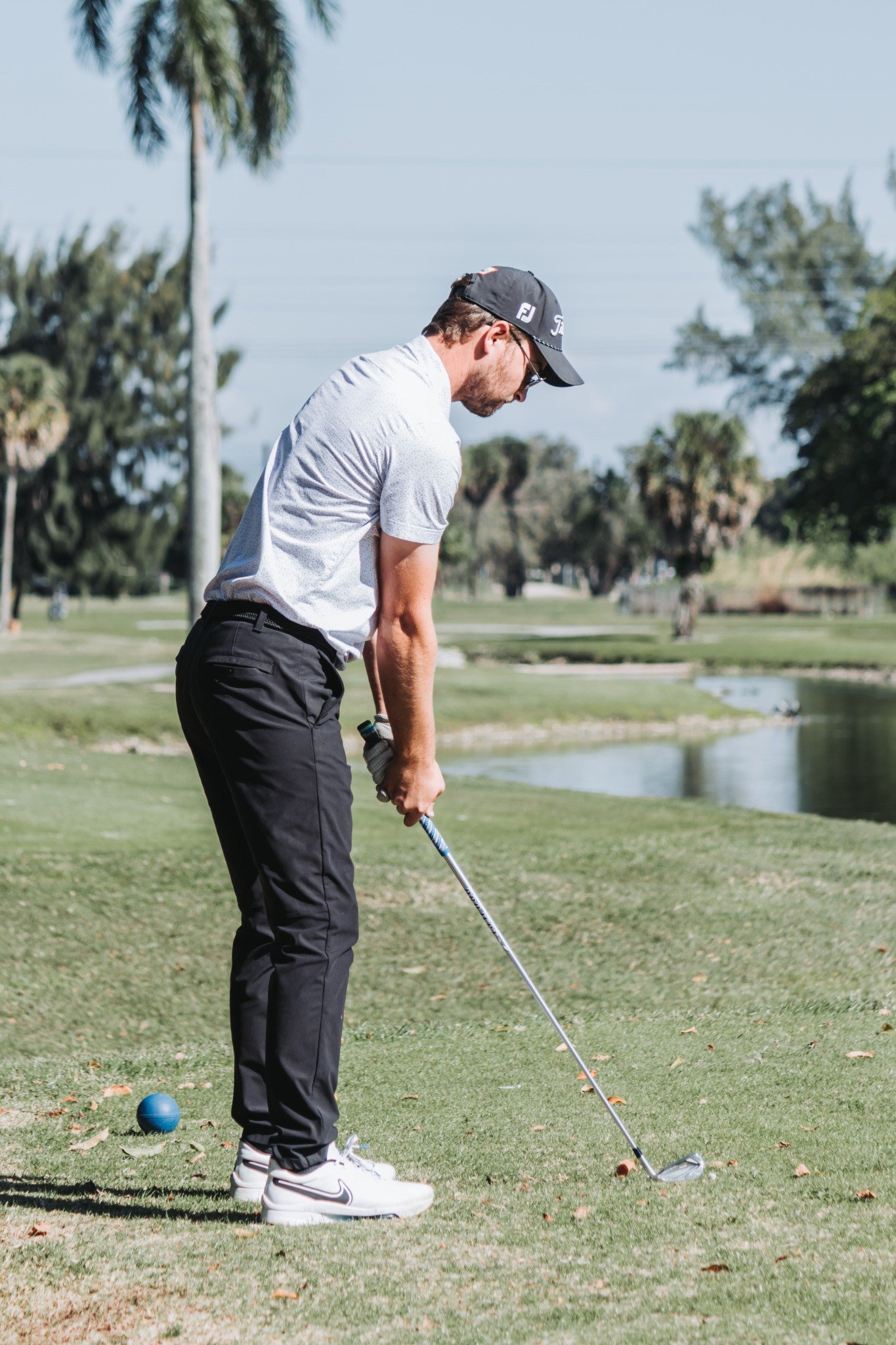 A man playing golf on a grassy course, preparing to hit a golf ball with a club, wearing a gray shirt, black pants, and a black cap, with a blue golf ball on the ground and a small water body in the background.
