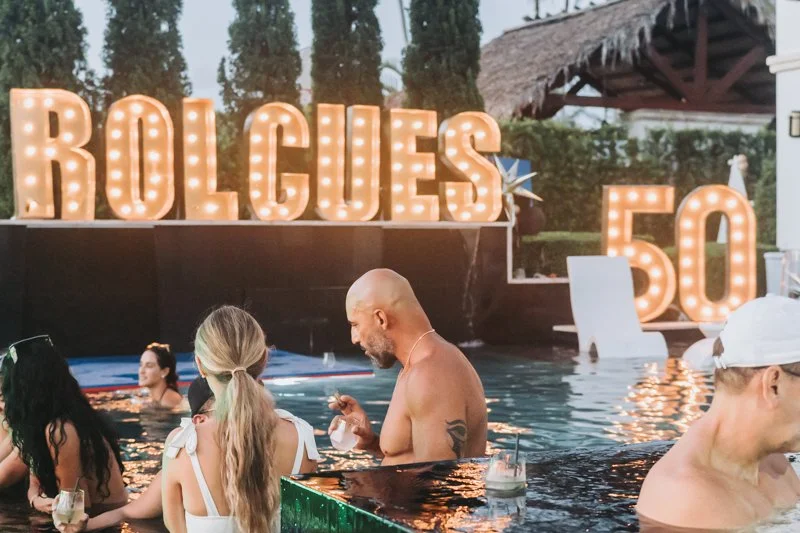 People in a pool with large illuminated marquee letters spelling 'ROLLES' and a sign with the number 50 in the background.